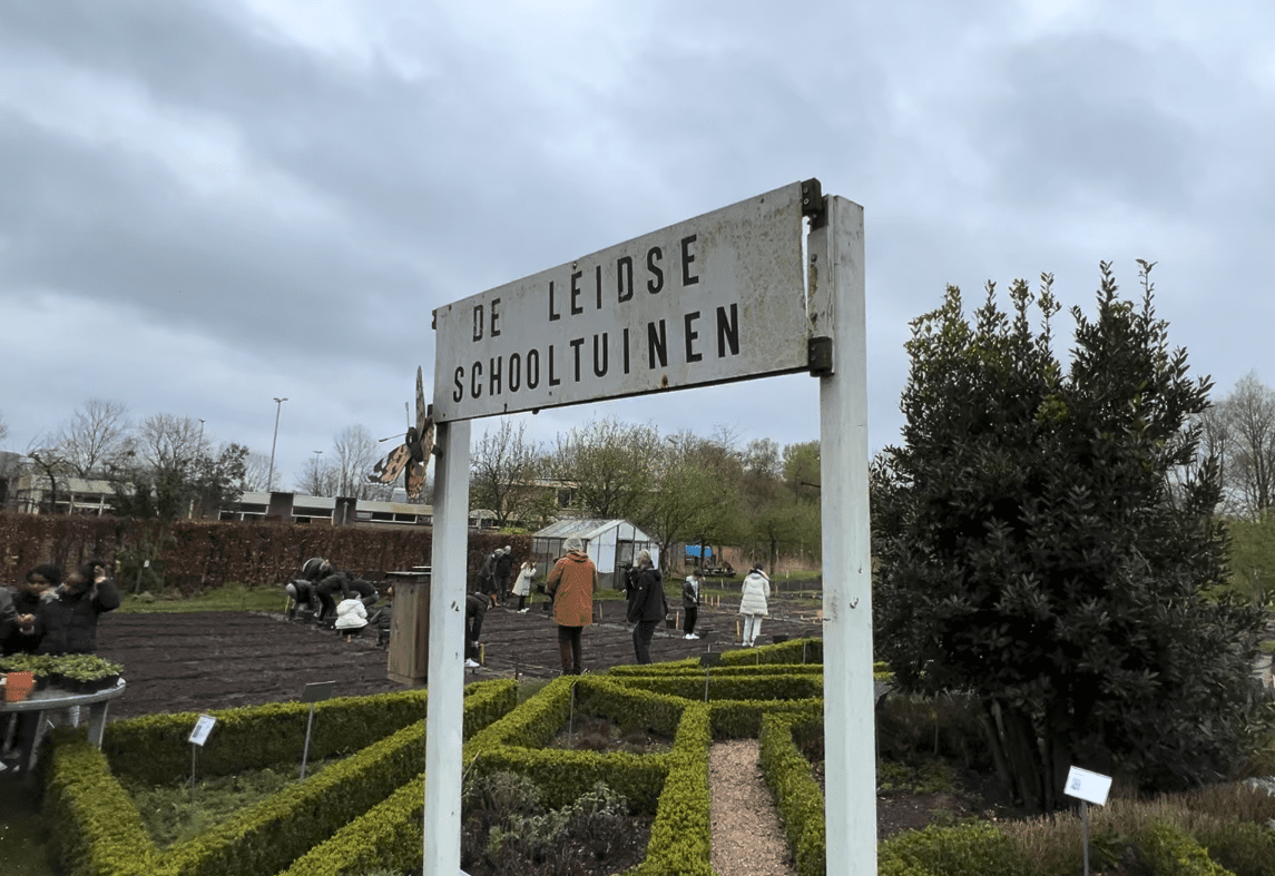 Bord "De Leidse Schooltuinen" met mensen die in een tuin werken op een bewolkte dag.