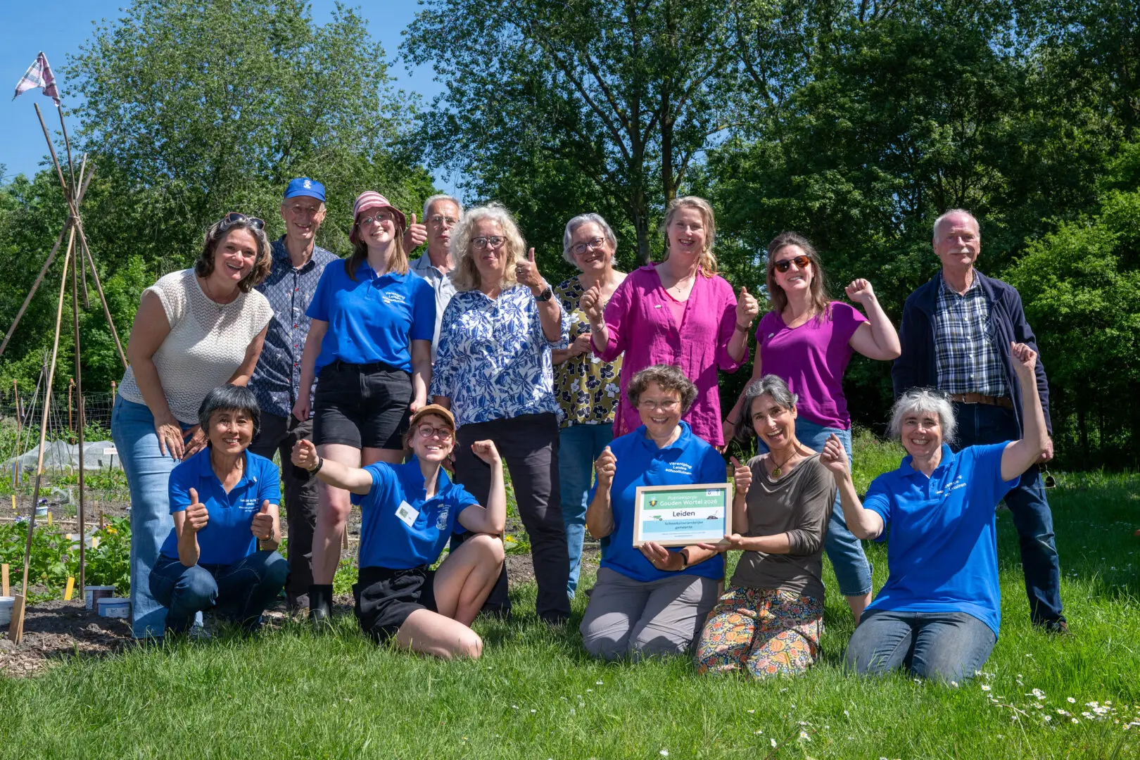 Groep mensen buiten in een tuin, sommigen in blauwe shirts, houden een oorkonde omhoog.
