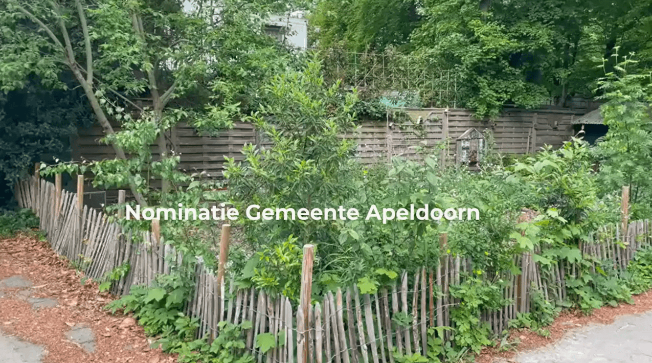 Stadstuin met houten hek, omgeven door bomen. Tekst: Nominatie Gemeente Apeldoorn.