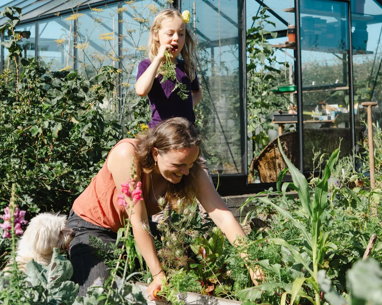Een vrouw en kind tuinieren met een hond bij een kas, omringd door groene planten.