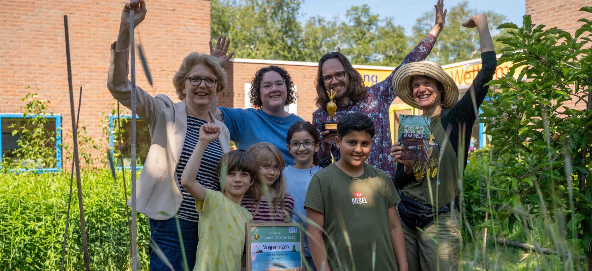 Groep mensen juichend in een tuin met een trofee en een plakkaat, zonnige dag op de achtergrond.